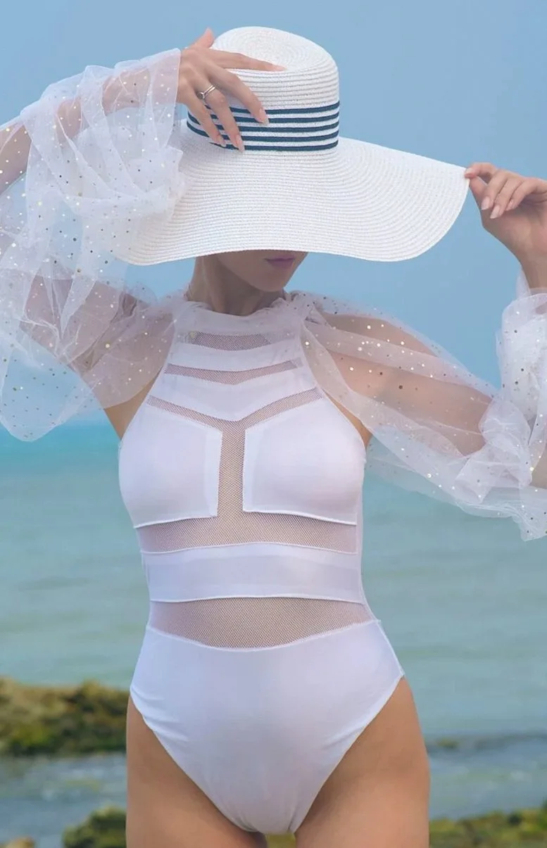 Woman in a white high-leg one-piece with sheer sleeves and a wide-brimmed hat by the sea