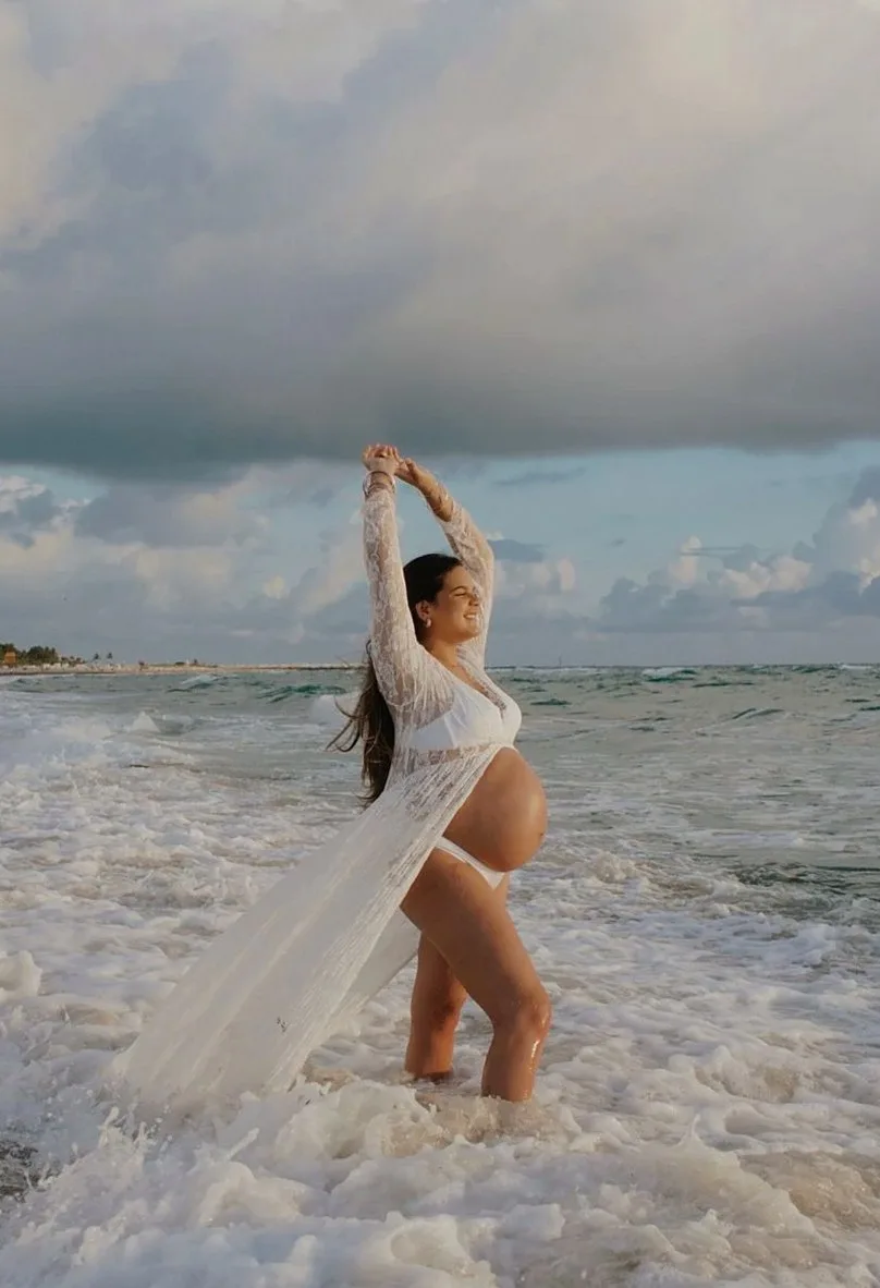 Woman in a long white cover-up standing in shallow surf with her arms raised under a cloudy sky