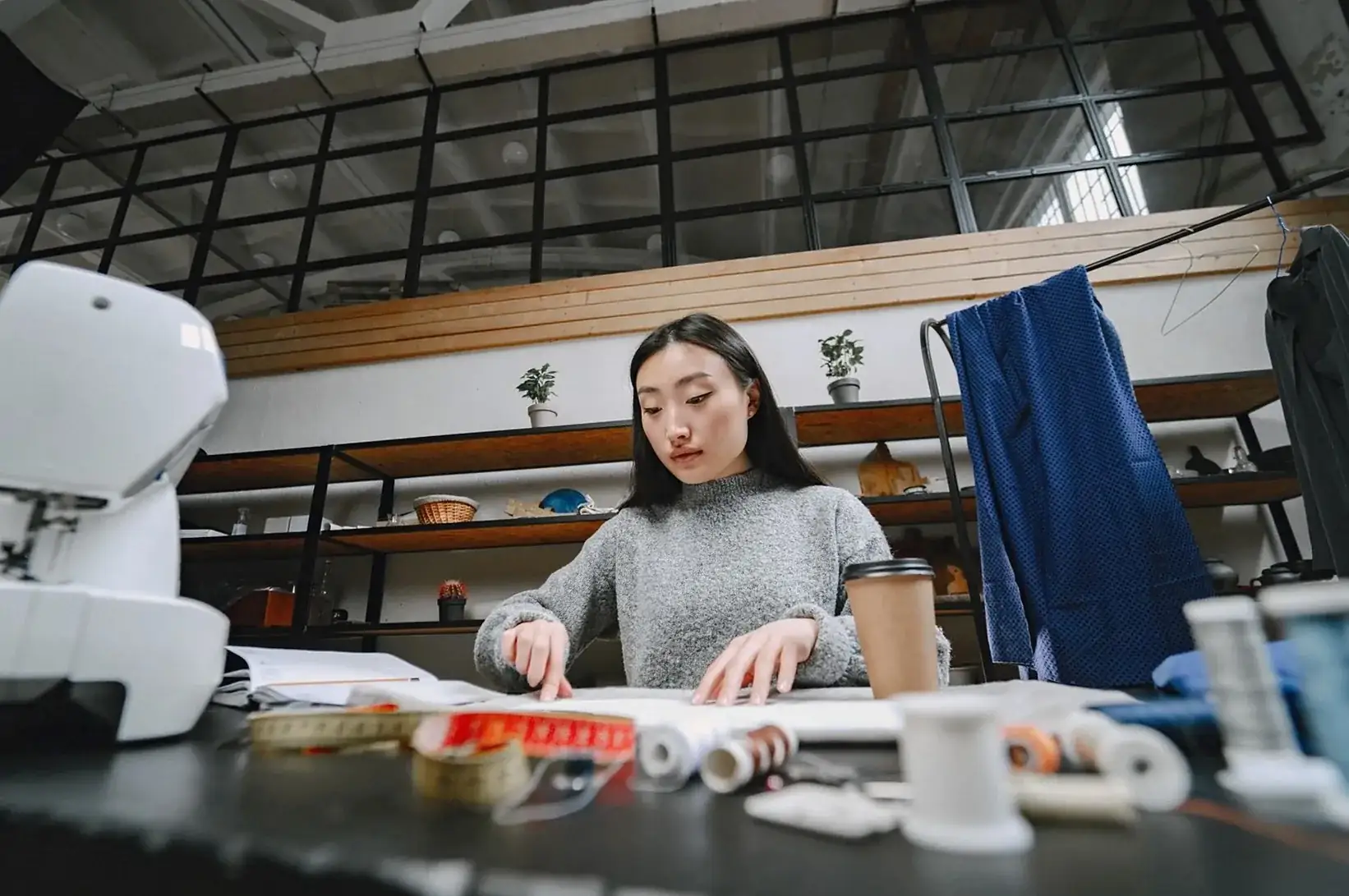 Woman working on a pattern at a studio table beside a sewing machine and rolls of fabric
